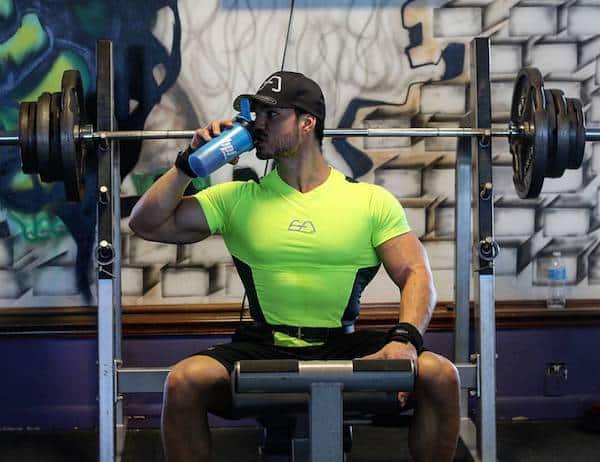 Le coach sportif Julien Quaglierini en train de consommer un shaker, assis sur un banc de musculation dans une salle de fitness.