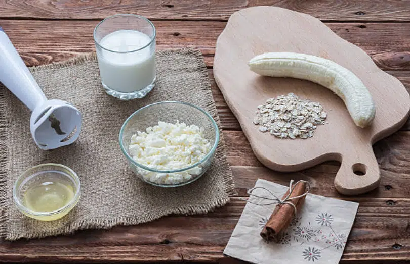 Une table avec différents ingrédients pour préparer un gainer maison : des flocons d'avoine, du lait, de la whey protein et de la banane.