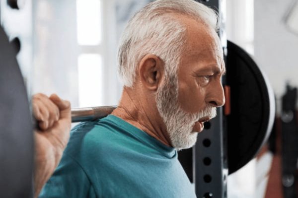 Un homme âgé, cheveux et barbe blancs, fait un exercice de musculation (squat) avec une barre sur les épaules.