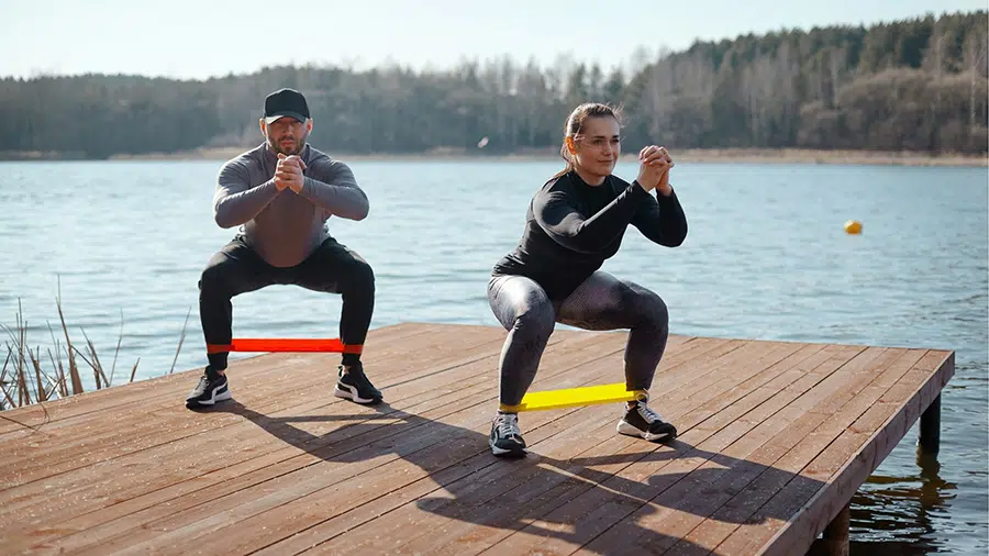 Au bord d'un lac, un homme et une femme font du squat avec élastique.