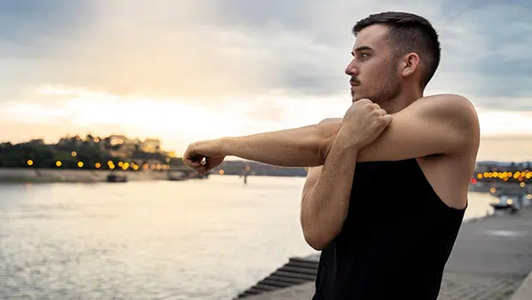 A man in a tank top performs a shoulder stretching exercise at the water's edge.