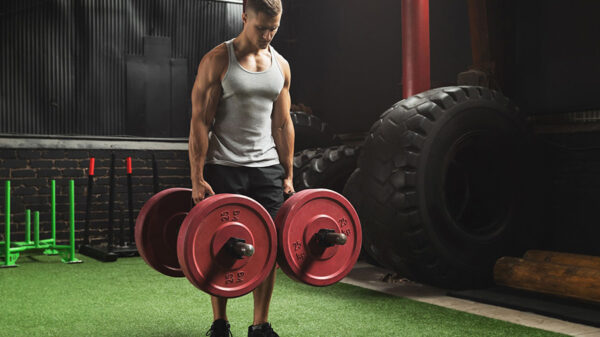 Un homme en débardeur effectue l'exercice du farmer walk dans une salle de musculation