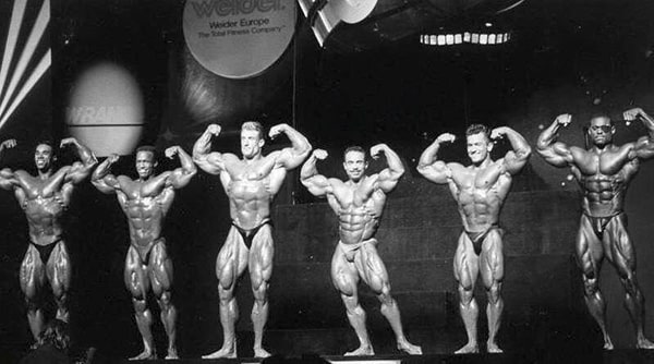 6 athletes are on stage during a bodybuilding contest, including Momo Benaziza, much smaller than the other competitors.