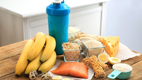 Une table avec de nombreux aliments : des bananes, du saumon, de la whey proteines, du fromage et un shaker pour les compléments alimentaires.