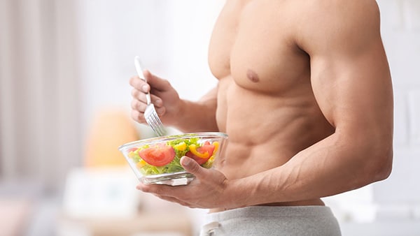 A shirtless muscular man eats a bodybuilding meal of salad, tomatoes and vegetables.