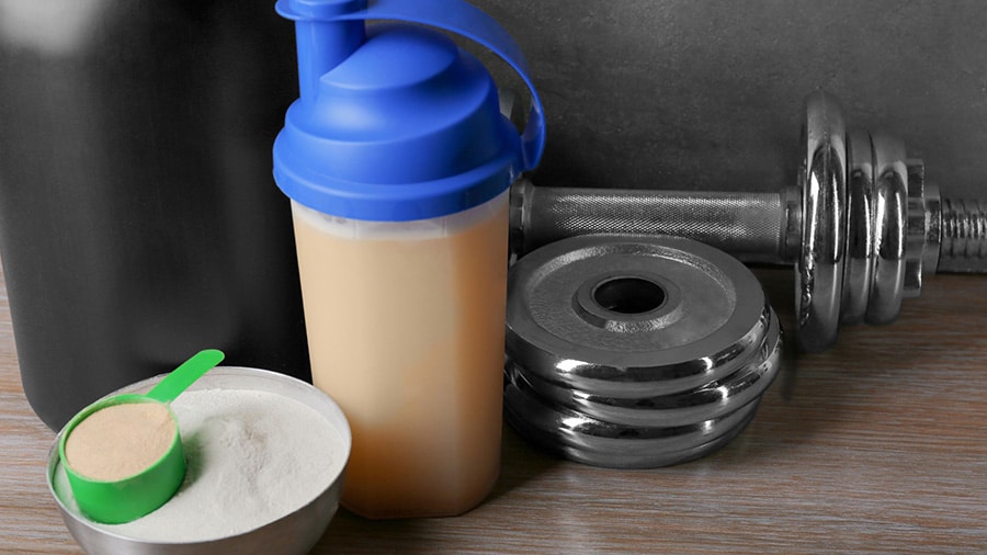 Un shaker composé d'un mass gainer, un bol avec de la poudre de protéines et un haltère de musculation.