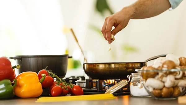 Un homme en cuisine ajoute des ingrédients dans une poêle, avec des légumes posés devant lui.