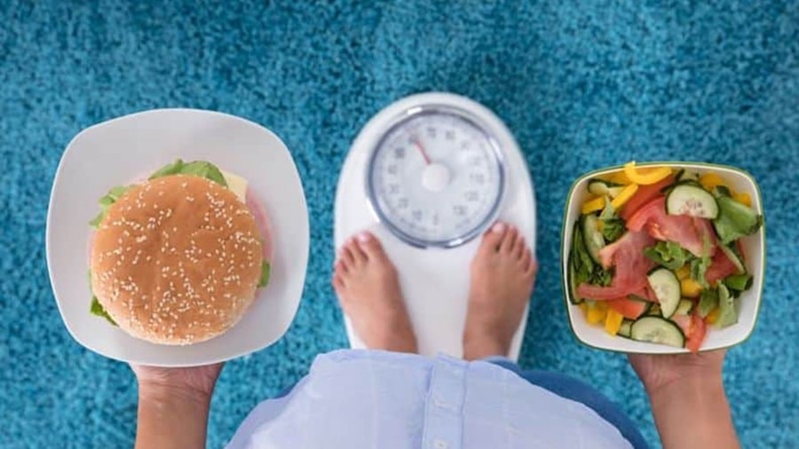 Le dilemme du rééquilibrage alimentaire : une personne debout sur une balance tient un hamburger dans sa main gauche et une assiette de légumes dans sa main droite.