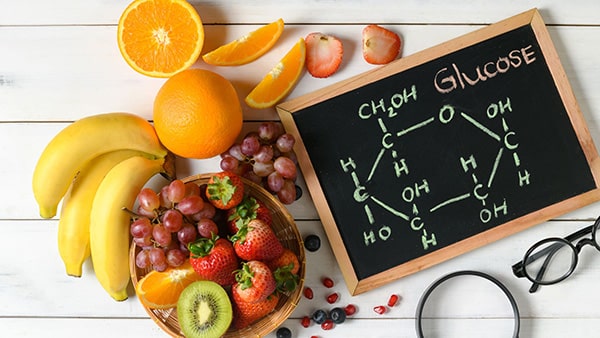 Une table avec des fruits et des légumes (banane, orange, fraise, kiwi) et un tableau noir représentant le glucose.