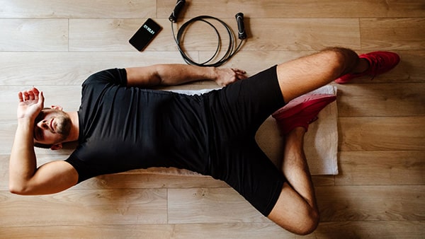 Un sportif est allongé sur une serviette posée sur le sol, avec un smartphone et une corde à sauter à côté de lui, visiblement fatigué par sa séance d'entraînement.