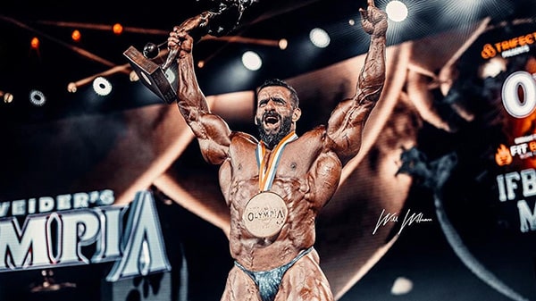 Hadi Choopan célèbre sa victoire à Mr. Olympia 2022, les bras en l'air, la médaille au tour du cou et le trophée dans sa main droite.