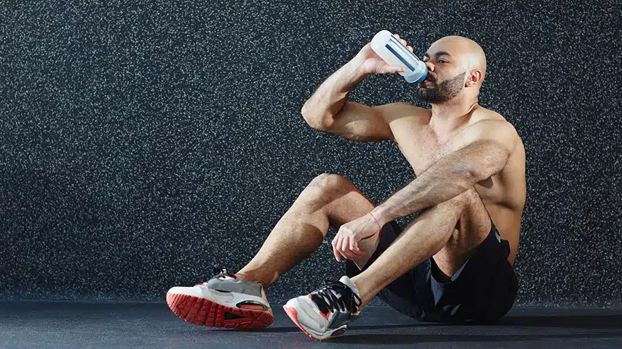 Un sportif torse nu, assis sur le sol, boit de l'eau dans une gourde.