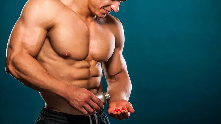 Un homme sportif, torse nu et musclé, verse dans sa main des gélules de ZMA, un complément alimentaire composé de zinc, de magnésium et de vitamine.