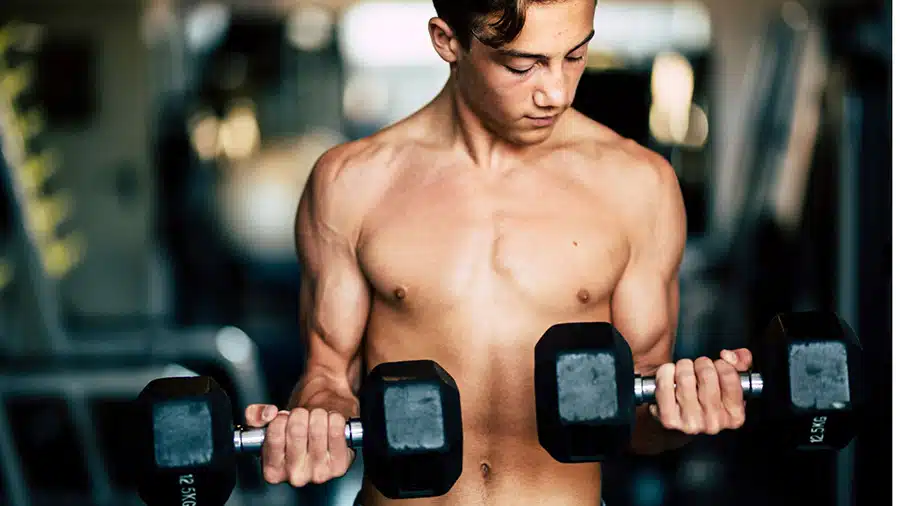Un jeune adolescent torse nu réalise un exercice de musculation avec des haltères dans une salle de sport.