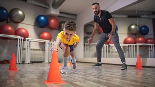 Un enfant effectue des exercices de fitness dans une salle de sport, enfreint par un coach sportif.