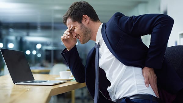 Un homme en costume-cravate, assis à un bureau devant un ordinateur, souffre de douleurs au bas du dos et se touche la région lombaire avec la main gauche.