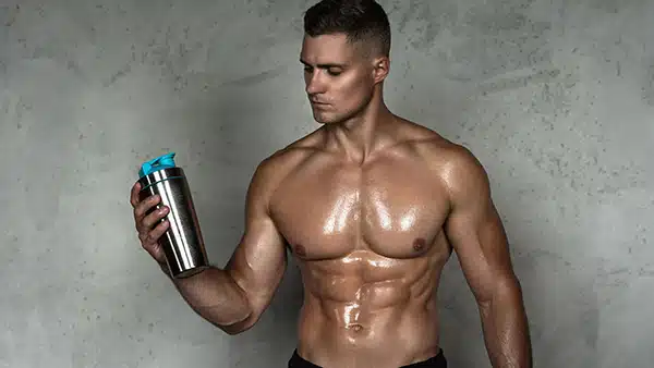 Un homme torse nu et musclé tient dans sa main droite un shaker de whey hydrolysée.