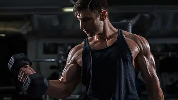 Un homme en débardeur effectue l'exercice du curl biceps dans une salle de musculation.