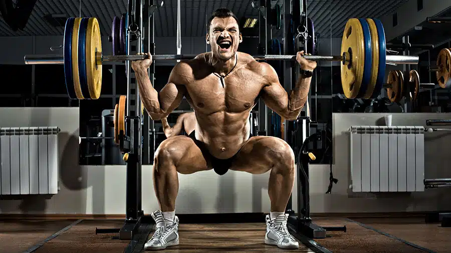 Un pratiquant de musculation torse nu effectue l'exercice du squat à la barre libre en appliquant la méthode pyramidale dans une salle de sport.