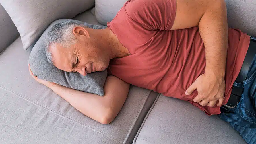 Un homme, vêtu d'un jean et d'un t-shirt rouge, est allongé sur un canapé et souffre de douleurs au ventre à cause de son périnée.
