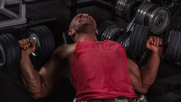Un sportif en débardeur rouge fait du développé couché aux haltères sur un banc de musculation pour muscler ses pectoraux.