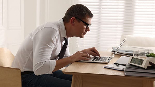 Un homme en chemise et en cravate travaille sur un ordinateur, assis à un bureau avec une mauvaise posture (dos creusé).