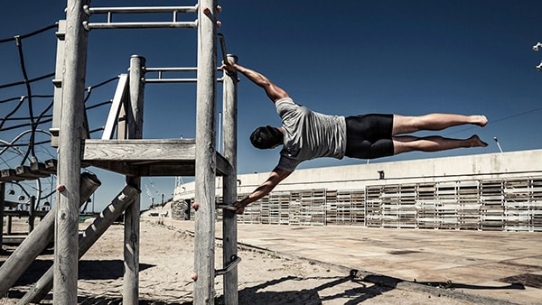 A man in t-shirt and shorts performs the human flag street workout on a structure at the beach.