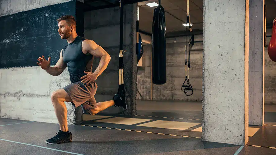Un homme en short et en débardeur effectue un exercice de fentes au TRX dans une salle de musculation pour renforcer ses muscles stabilisateurs.
