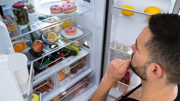 Un homme s'interroge sur ce qu'il va manger devant un frigo ouvert contenant des fruits et des donuts.