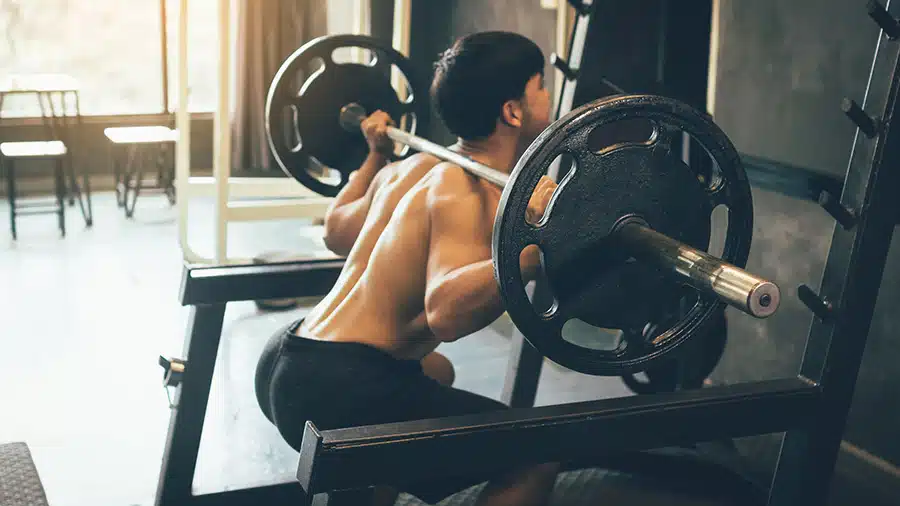 Un homme torse nu dans une salle de sport applique la méthode stato-dynamique sur l'exercice du squat à la barre.