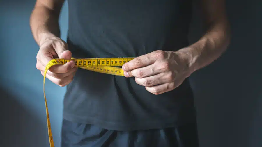 Un homme mesure son tour de ventre avec un mètre ruban pour suivre l'efficacité de sa recomposition corporelle.