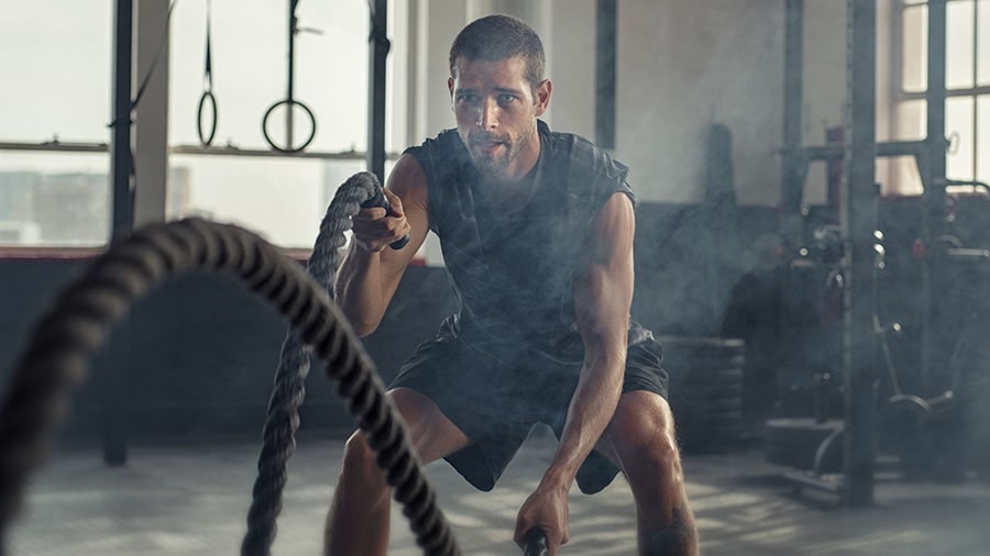 A man in a tank top in a gym works out with an undulating rope.