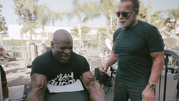 Bodybuilder Ronnie Coleman performs a biceps curl exercise, with Arnold Schwarzenegger cheering him on.
