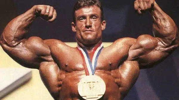 Le bodybuilder Dorian Yates torse nu, les muscles des biceps contractés, portant une médaille autour du cou.