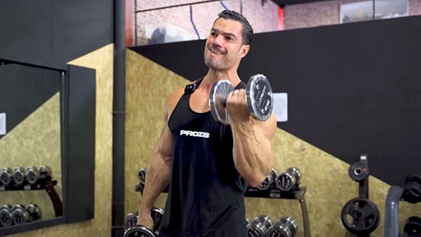 Sports coach Julien Quaglierini, wearing a black tank top, performs a biceps curl exercise in a weight room.