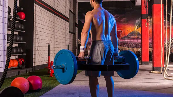 Un homme torse nu, de dos, tient une hex bar avec ses deux mains dans une salle de fitness.
