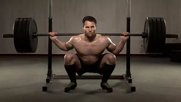 Un homme torse nu fait du squat, barre de musculation sur les épaules, dans une salle de fitness.