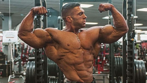 Professional bodybuilder Derek Lunsford, Mr Olympia 2023, shirtless in a weight room performing a double biceps pose.