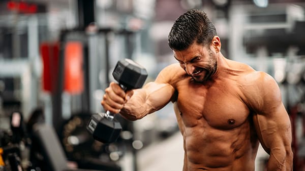 Un homme torse nu dans une salle de fitness en train de faire un exercice de musculation (élévations frontales) pour les épaules avec un haltère dans la main droite.
