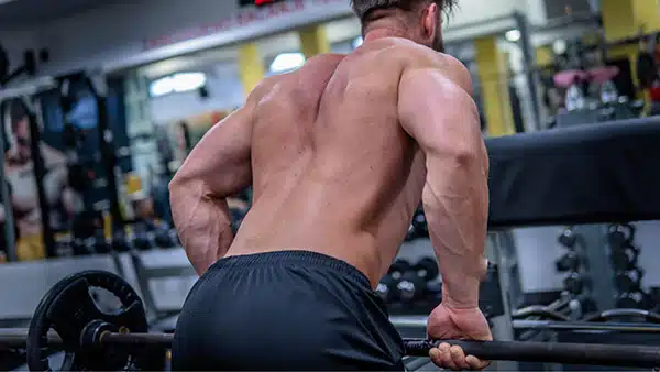 Dans une salle de musculation, un homme torse nu, de dos, effectue un exercice pour développer le dos avec une barre.