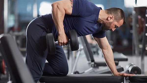 Dans une salle de sport, un homme réalise l'exercice du tirage bûcheron sur un banc de musculation, avec un haltère dans la main gauche.