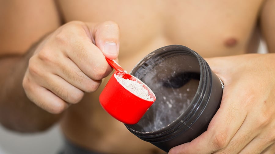 A shirtless man pours a dose of vegan whey into a shaker.