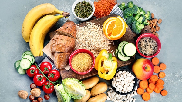 Un ensemble d'aliments posés sur une table : des bananes, des tomates, des brocolis, du pain, du riz, de la carotte, des pommes de terre, du poivron, des amandes, etc.