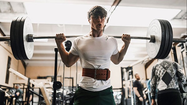 L'influenceur fitness David Laid en pleine séance de musculation dans une salle de sport, en train de faire une squat avec une barre sur les épaules et une ceinture autour de la taille.