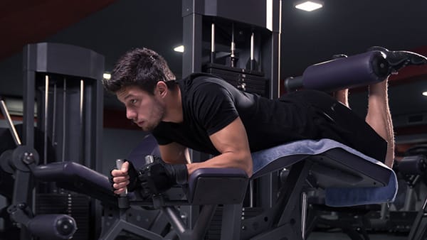 Un homme habillé en noir fait du leg curl allongé sur une machine de musculation dans une salle de sport pour renforcer ses ischio-jambiers.