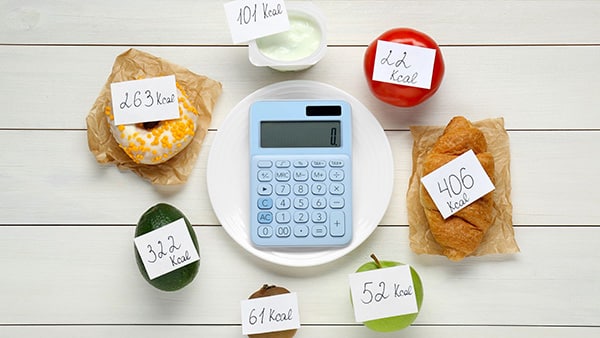 Une calculette dans une assiette, entourée d'aliments (pomme, croissant, tomate, kiwi, avocat, donut) avec, pour chacun d'entre eux, la correspondance en calories.