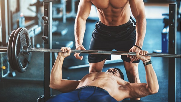 2 bare-chested athletes doing bench presses in a weight room, the first ensuring the safety of the second.