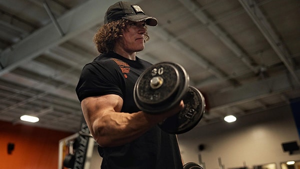 Le bodybuilder Sam Sulek réalise un exercice de musculation, le curl biceps, avec un haltère dans la main droite.