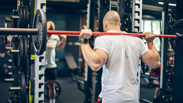 Un homme de dos en t-shirt blanc fait du squat, barre de musculation sur les épaules, dans une salle de sport.
