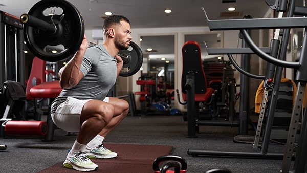A man in shorts does squats with a barbell in a fitness center.